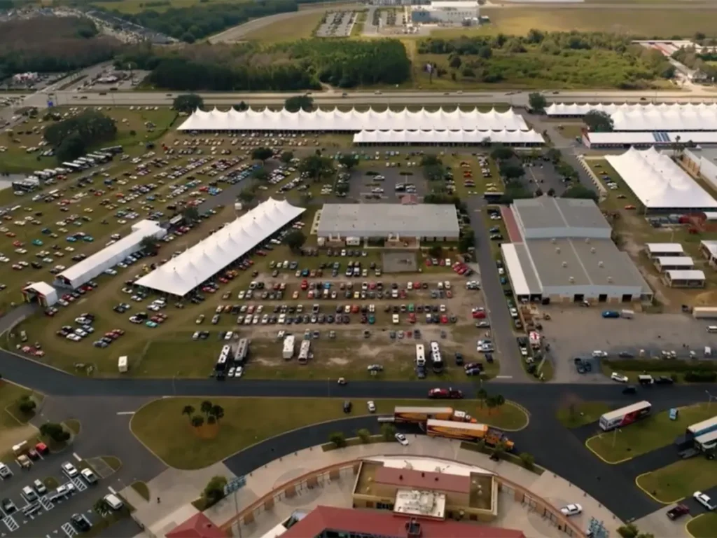 A high aerial view showing a vast outdoor event space dominated by large white temporary tents and buildings. Numerous classic cars are parked in rows across grassy fields and paved lots. Curved roads weave through the complex, with some large transport trucks visible. Distant urban areas and woodlands frame the horizon under a partly cloudy sky.