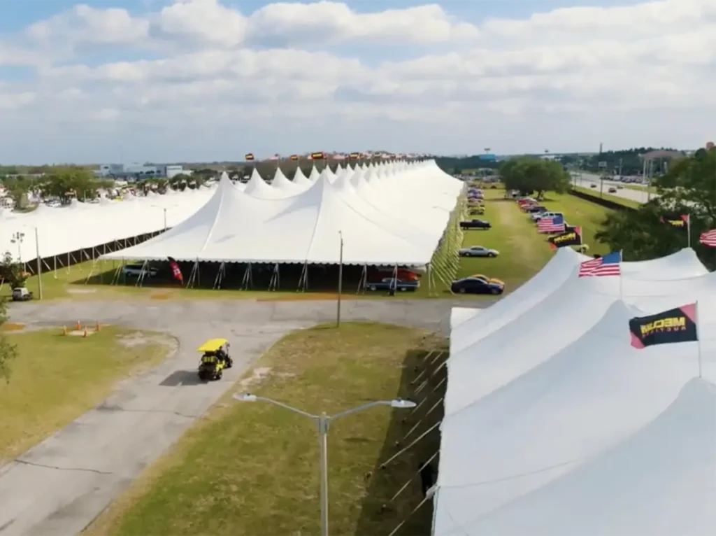 An aerial view showing a long row of large white tension pole tents with distinct peaked roofs stretching into the distance. Cars are parked neatly alongside the tents. A yellow golf cart is driving on a paved path in the foreground. Trees and other event structures are visible, with American flags and Mecum Auctions banners displayed on some of the tents, all under a blue sky with scattered clouds.