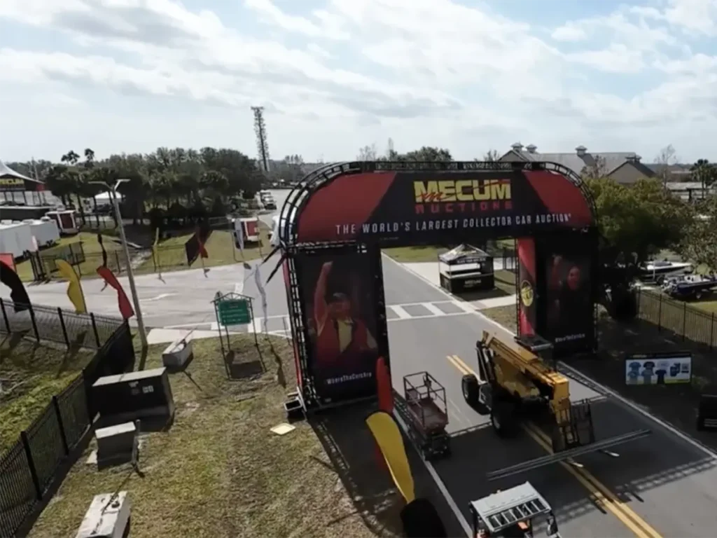 A large red and black archway with Mecum Auctions The World's Largest Collector Car Auction stands over a paved road. Beneath the arch, a yellow forklift is moving equipment. Various banners and flags line the road, which appears to be an entrance to an event. In the background, other event structures and trees are visible under a bright, partly cloudy sky.