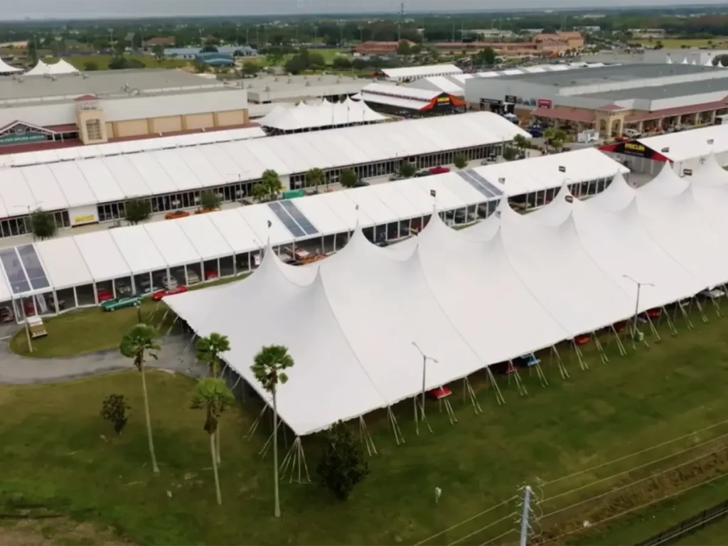 An expansive aerial view of a vast complex of large white temporary structures and tents, some with visible solar panels on their roofs. Numerous classic cars are parked beneath and around the tents. Green grass with palm trees lines the foreground, and other buildings and urban development are visible in the distance under a cloudy sky.