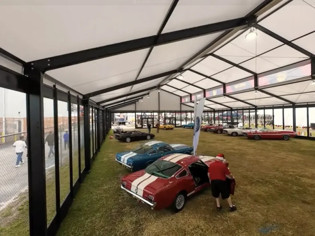 The interior of a large temporary structure with a white fabric roof and black metal framework. Numerous classic cars, including a red Ford Mustang with white racing stripes, are displayed on a lush green lawn. People are visible near the cars, and the side of the tent features clear, window-like panels that look out onto an outdoor area.