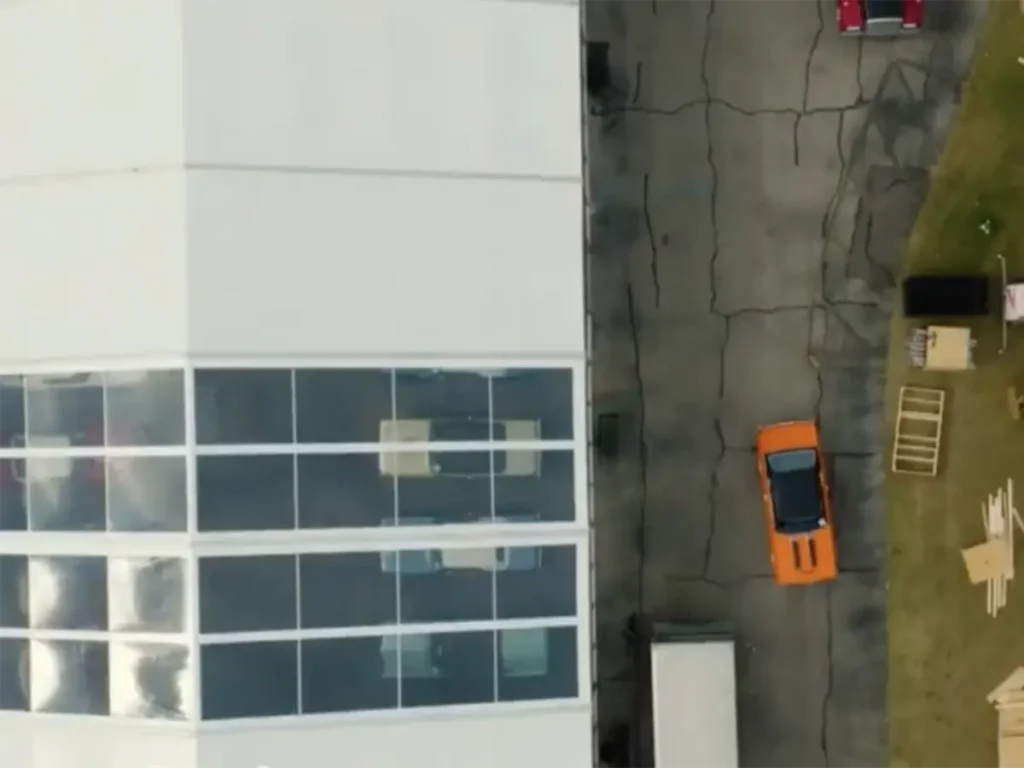 An overhead aerial view of a large, modern, white temporary structure with reflective windows on the left. To the right is an asphalt road with visible cracks, an orange sports car, and a red vehicle further down. Beside the road is a grassy strip and an area with various wooden crates, pallets, and equipment, indicating that an event is either being set up or broken down. A white truck trailer is also visible on the far right.