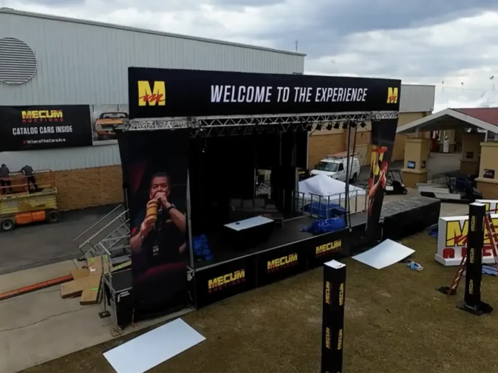 An outdoor stage setup with large black banners featuring Mecum Auctions and WELCOME TO THE EXPERIENCE. The stage is adorned with a variety of lights and equipment. To the left, a yellow lift is positioned near a white building with a Mecum Auctions Catalog Cars Inside banner. In the background, other temporary structures and buildings are visible under a partly cloudy sky.