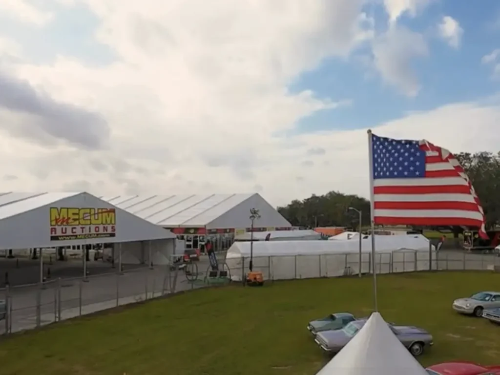 An outdoor view of several large white temporary structures, likely tents, at an event. The structure in the foreground has a Mecum Auctions banner. A large American flag proudly waves from a flagpole in the middle ground. Several classic cars are parked on the green grass, and trucks are visible in the background under a partly cloudy sky.