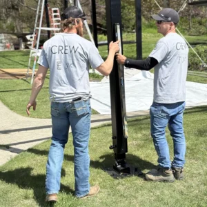 Two JK Rentals crew leaders align a vertical tent support using a level during a sunny outdoor setup. Both wear matching grey crew t-shirts and jeans.