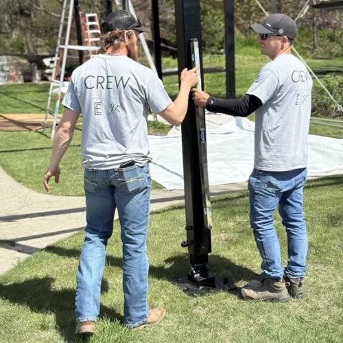 Two JK Rentals crew leaders align a vertical tent support using a level during a sunny outdoor setup. Both wear matching grey crew t-shirts and jeans.