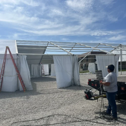 JK Rentals field worker guides a tensioned tent top into place using ropes while standing near a truck and several ladders.