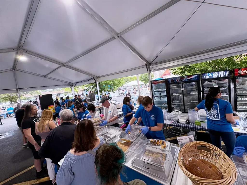 Volunteers in blue shirts serve trays of traditional Greek pastries under a white tent. Floyeres and other sweets are displayed in clear containers on checkered tablecloths beside a commercial fridge.