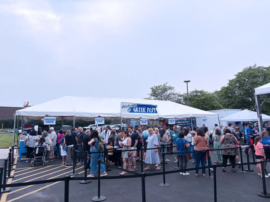 Crowds line up at the food tent labeled “Welcome to Greek Fest” with separate areas for ordering and pickup. Signage and crowd control barriers organize the lines, and tents extend into the background.