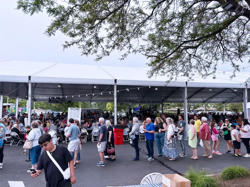 Street-level view of a long line of people waiting outside a large open-side tent at Greek Fest with signage reading 'OPA! WE’RE ALL GREEK THIS WEEKEND.'