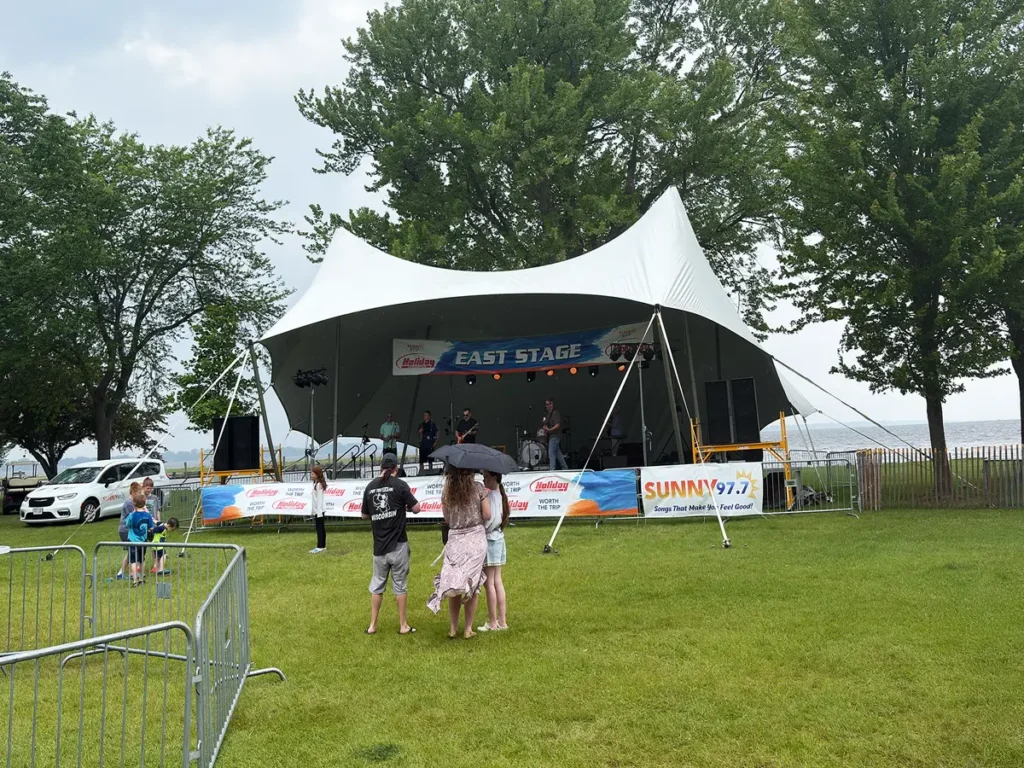 Bandshell-style tension tent used for an outdoor concert stage with performers and spectators under cloudy skies