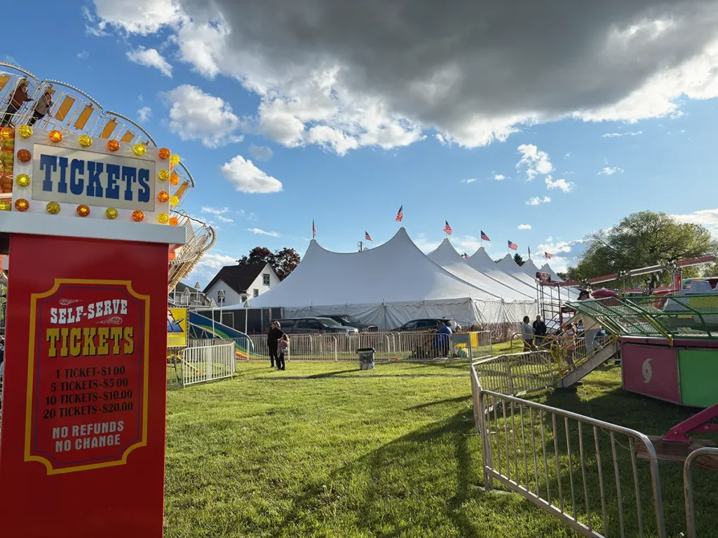 Tension tent installed at a community carnival with flags, ticket booth, and amusement rides in view