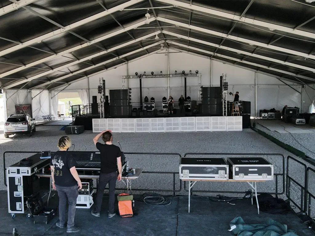 Stage being constructed under a large black clearspan tent with sound equipment visible.