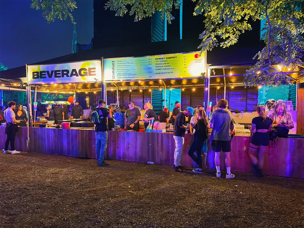 Beverage tents lit with string lights at night, with green beams from the nearby stage cutting across the sky.