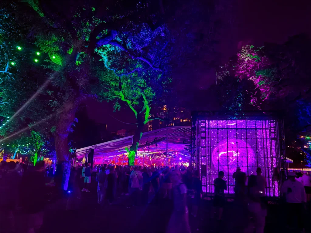 “The Oracle” art installation by The Vessel glowing in the trees at ARC Music Festival, with the Arcum clearspan structure illuminated in the background.