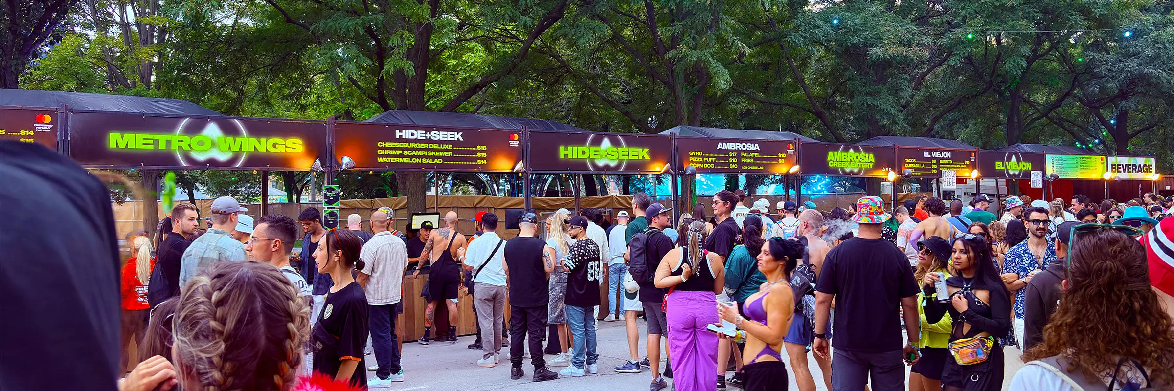 Wide view of the ARC Music Festival food vendor row, with multiple tents, neon menu boards and crowds moving along the pathway.