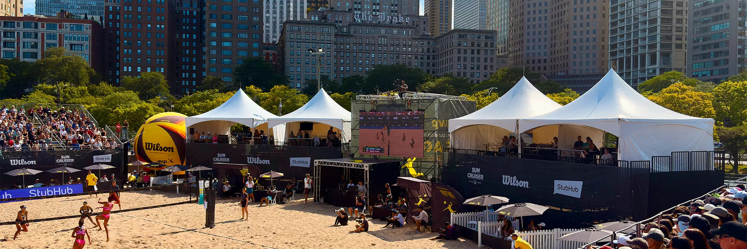 Panoramic view of the AVP main court with players, VIP tents and elevated spectator platforms.