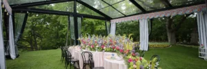 Pastel floral arrangements lining round tables beneath a black-frame clear tent set on a green lawn.