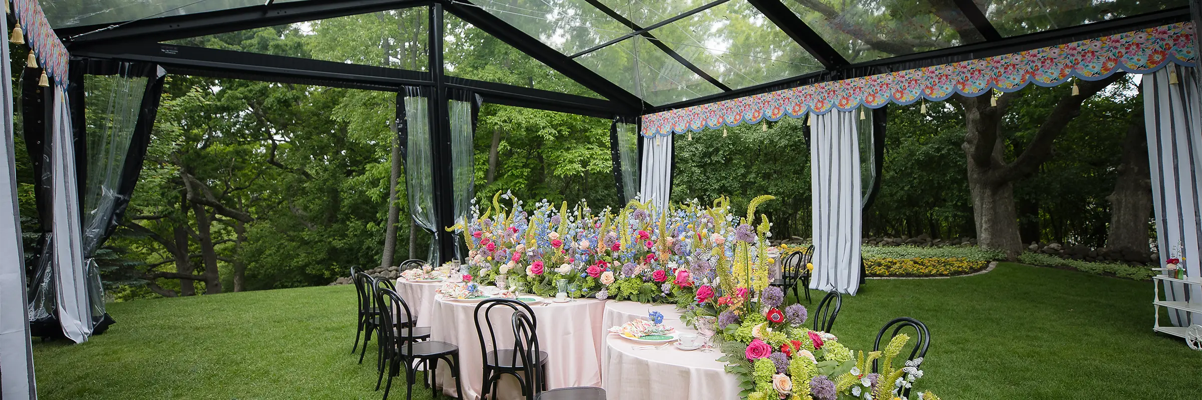 Pastel floral arrangements lining round tables beneath a black-frame clear tent set on a green lawn.