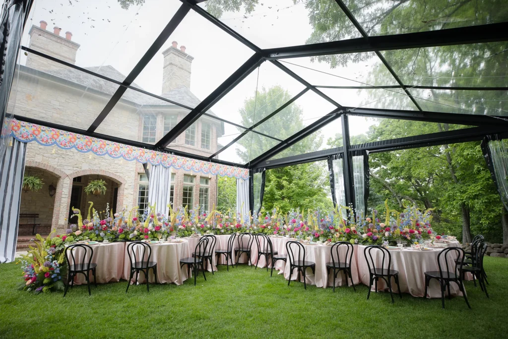 Elegant baby shower setup beneath a clear tent with pastel florals and a stone residence in the background.