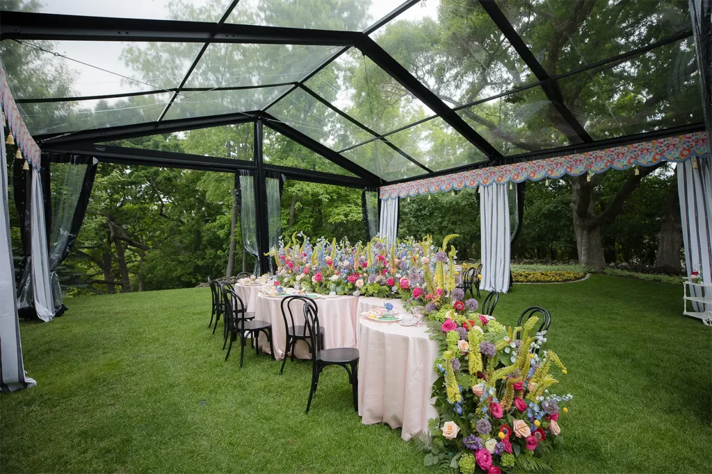 Colorful floral tablescape beneath a clear tent with open garden views and striped draping.