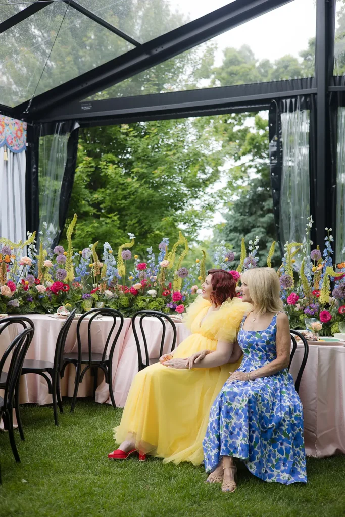 Two guests sit inside a clearspan tent surrounded by bright floral centerpieces and pastel table décor at a baby shower.