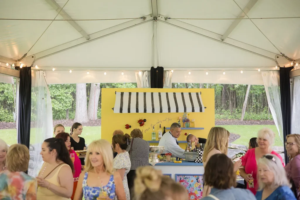 Guests mingle around a colorful bar setup under a frame tent with string lights at a baby shower.