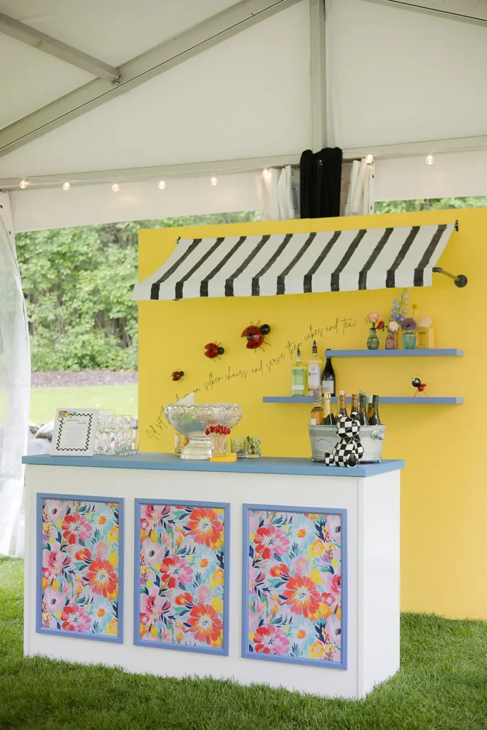 Floral-themed baby shower bar setup with a yellow backdrop, black-and-white striped awning, ladybug accents, and colorful floral panels under a white tent.