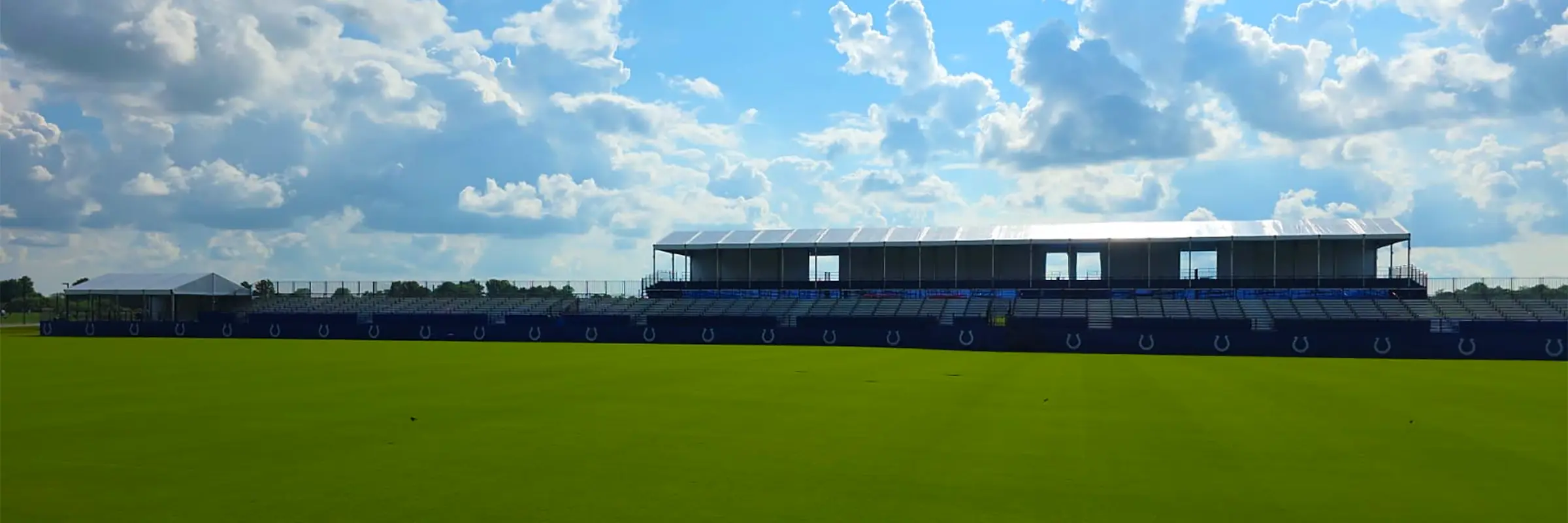 Large elevated grandstand with a clearspan structure roof at Colts Training Camp, viewed across the practice field under a bright sky.