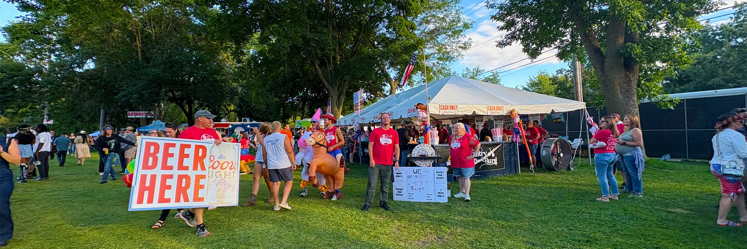 Volunteers working at a beer tent under a white frame tent at Country in the Burg, with a large “Beer Here” sign and festival guests nearby.