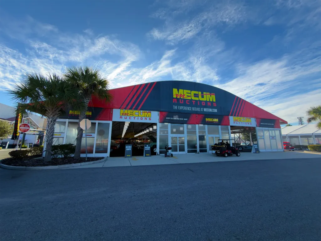 Front elevation of a JK Rentals clearspan with glass entry doors and Mecum Auctions branding, set among palm trees in Kissimmee, Florida.