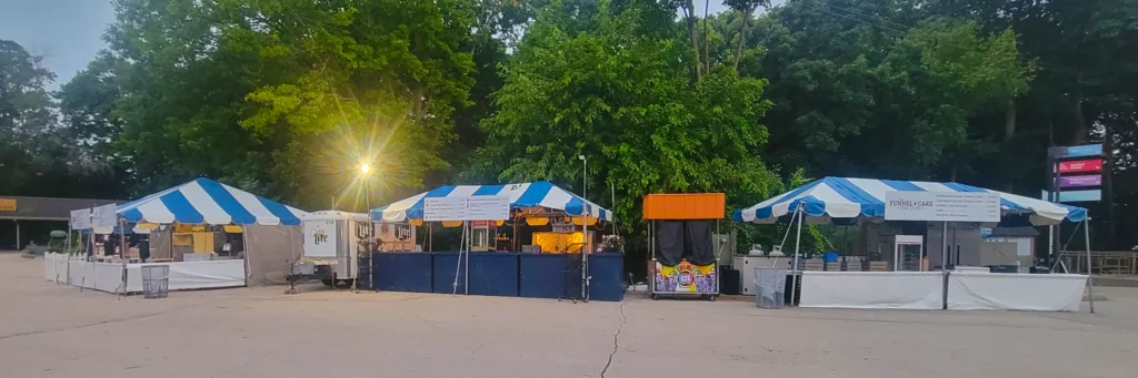 Wide view of multiple blue and white striped frame tent vendor booths set up as a food court area in the evening at Zoo A La Carte, Milwaukee County Zoo.