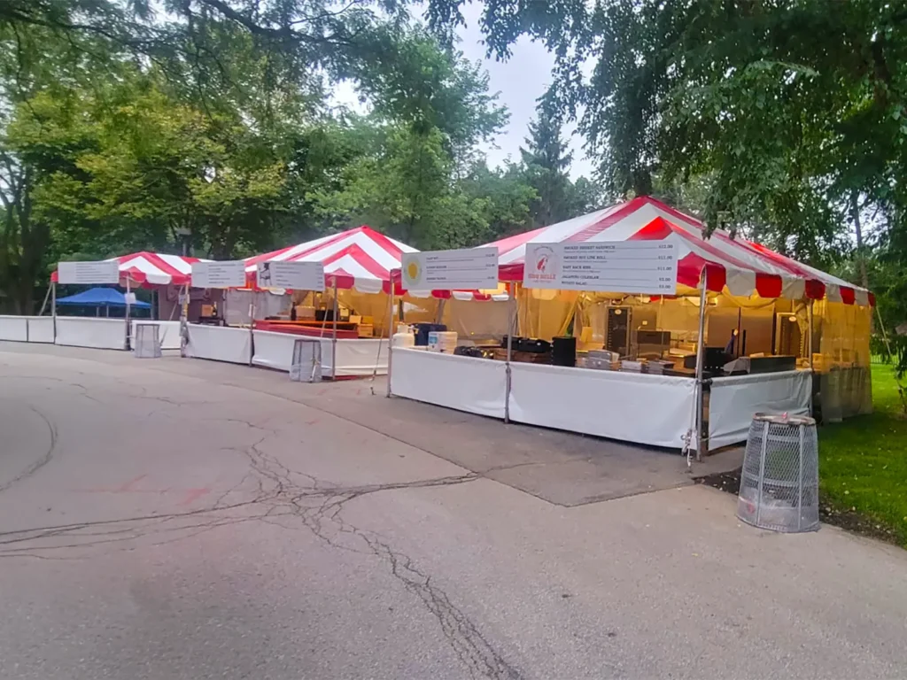 Long row of red and white striped frame tent vendor booths with menu boards and interior lighting along the zoo path during Zoo A La Carte.