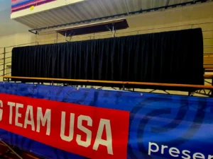 Black pipe and drape enclosure used to conceal an elevated platform inside an indoor sports venue.
