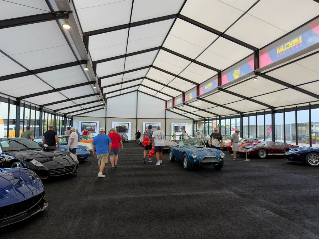 Interior of a clearspan structure at Mecum Auto Auctions with classic cars on display and visitors walking through a wide center aisle.