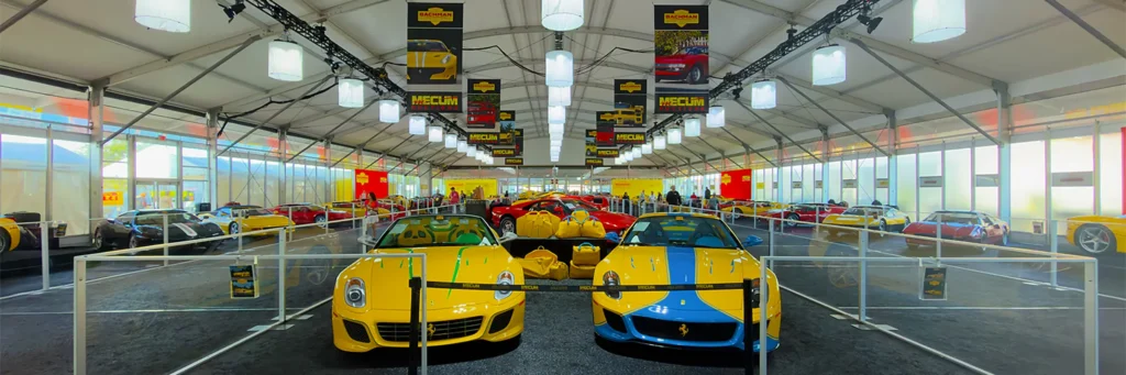 Wide interior view of a Mecum auto auction display tent with classic cars, overhead lighting, and crowd-control railings.