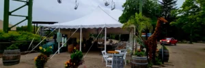 White high-peak pole tent seating area with cocktail tables, barrel accents, and a giraffe feature near the pathway at Zoo A La Carte in Milwaukee County Zoo.