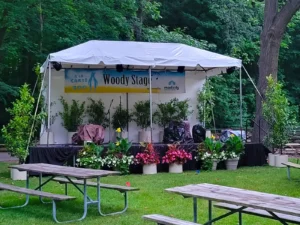 White canopy stage labeled Woody Stage with plants and floral décor in front, set on grass for live entertainment at Zoo A La Carte in Milwaukee County Zoo.