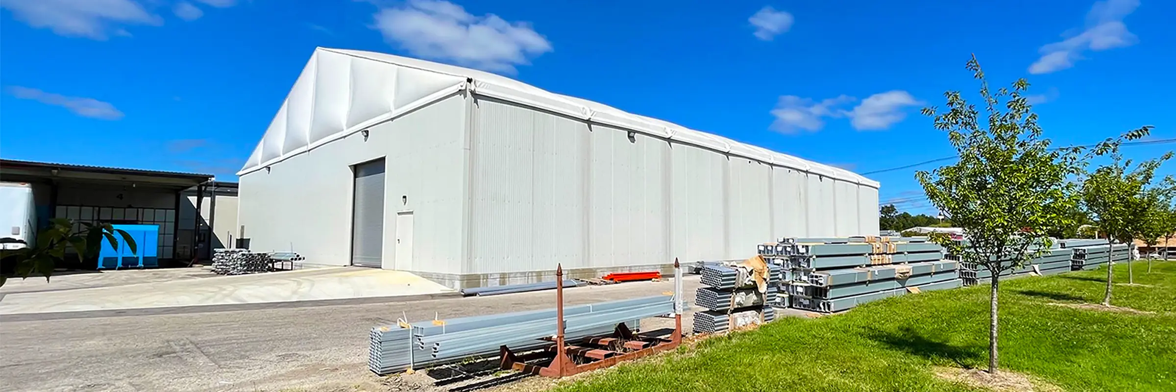 Wide panoramic view of a hard-wall clearspan warehouse exterior with stacked beams and panels along the drive lane.