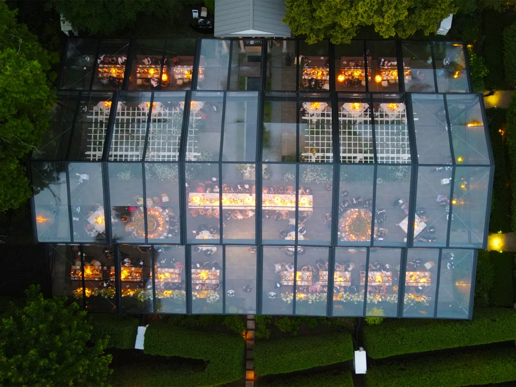 Aerial view of a JK Rentals Levo clearspan tent with transparent roof panels, glowing reception tables, dance floor, and wedding guests gathered outdoors at night.