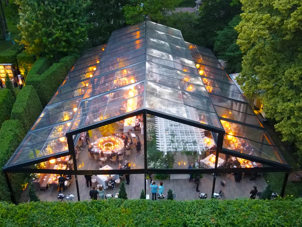 Aerial view of a JK Rentals Levo clearspan wedding tent with clear black vinyl panels, warm reception lighting, guest seating, floral decor, and a garden-style event layout.